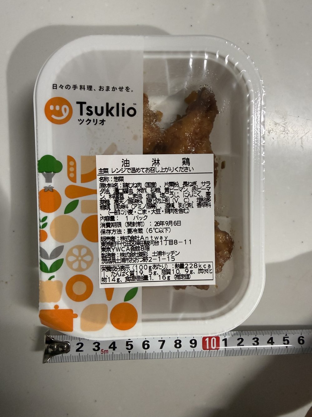 油淋鶏のパック。原材料名・栄養成分表示・消費期限・製造者情報がラベルに記載され、容器の横幅をメジャーで測っている